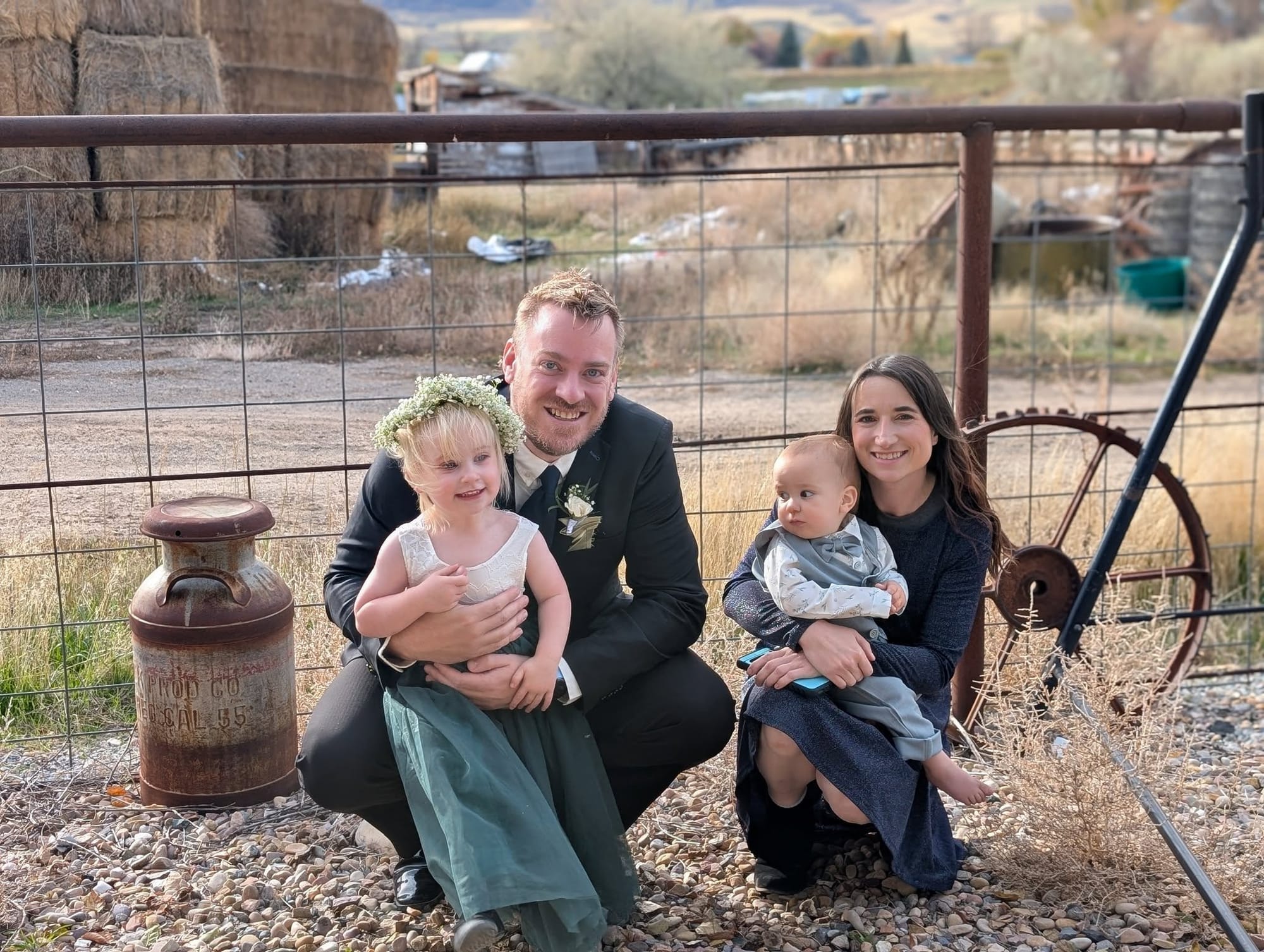A smiling family of four posing outdoors in front of a rustic fence, with the father in a suit kneeling beside a blonde toddler in a flower crown and green dress, and the mother sitting beside them holding their baby boy.