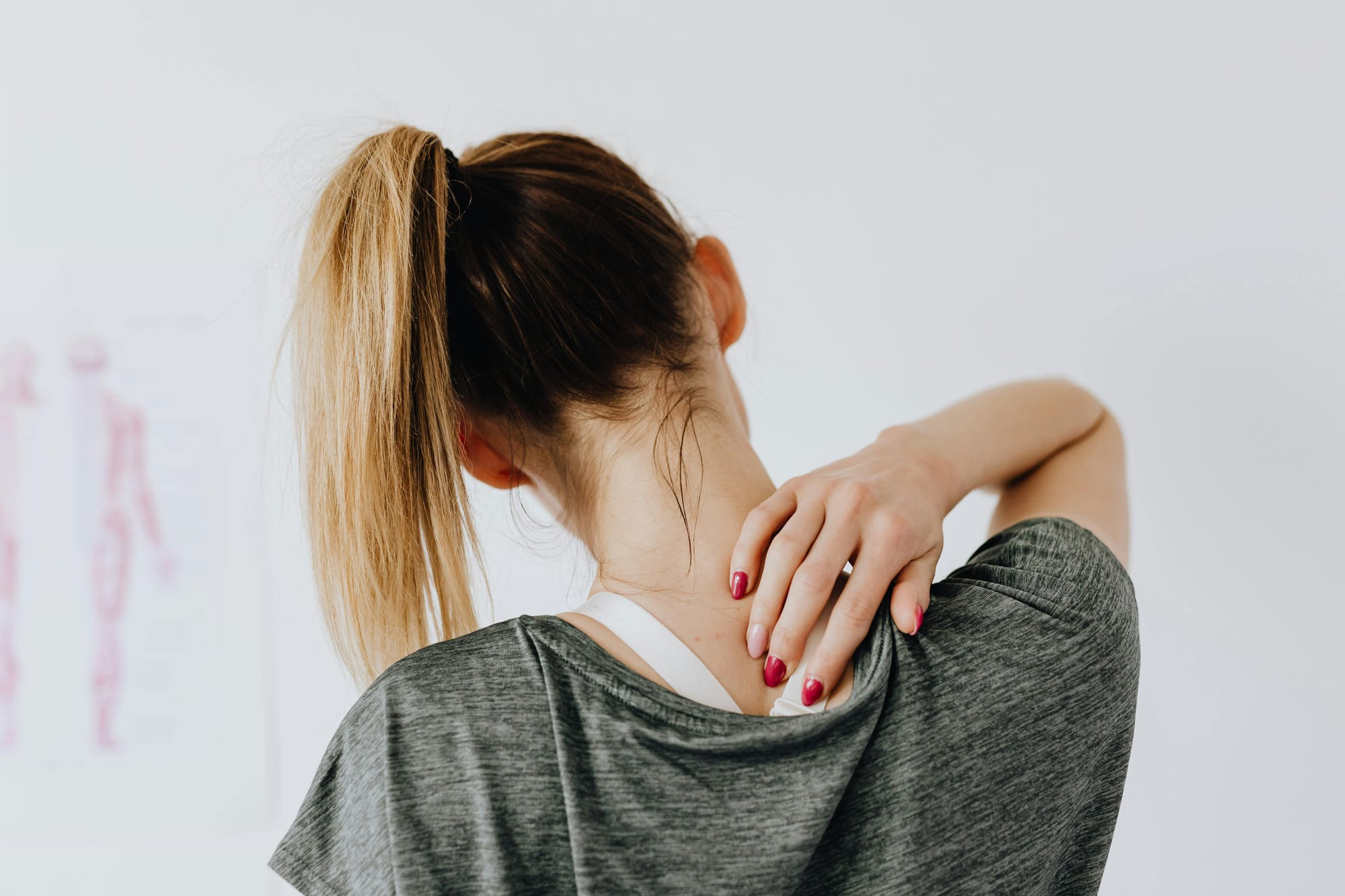 A woman with a blonde ponytail touching the back of her neck as if experiencing shoulder or neck tension.