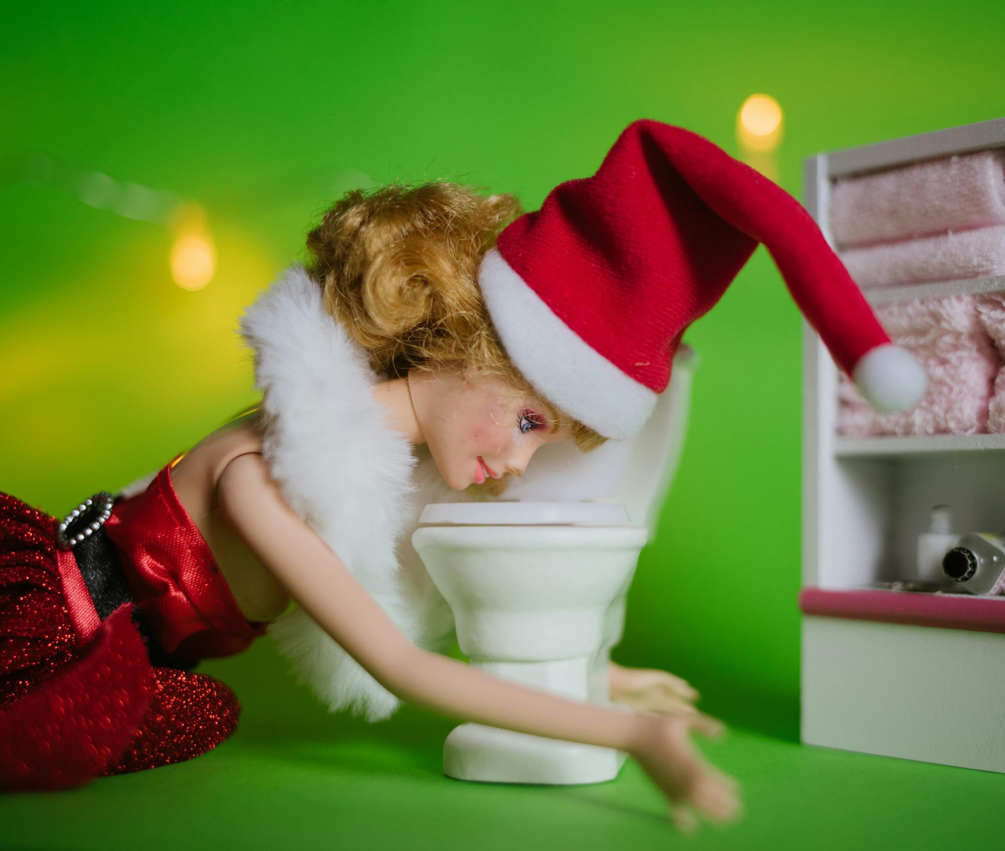 A humorous scene of a Barbie doll dressed in a red holiday outfit and Santa hat leaning over a miniature toilet as if sick.