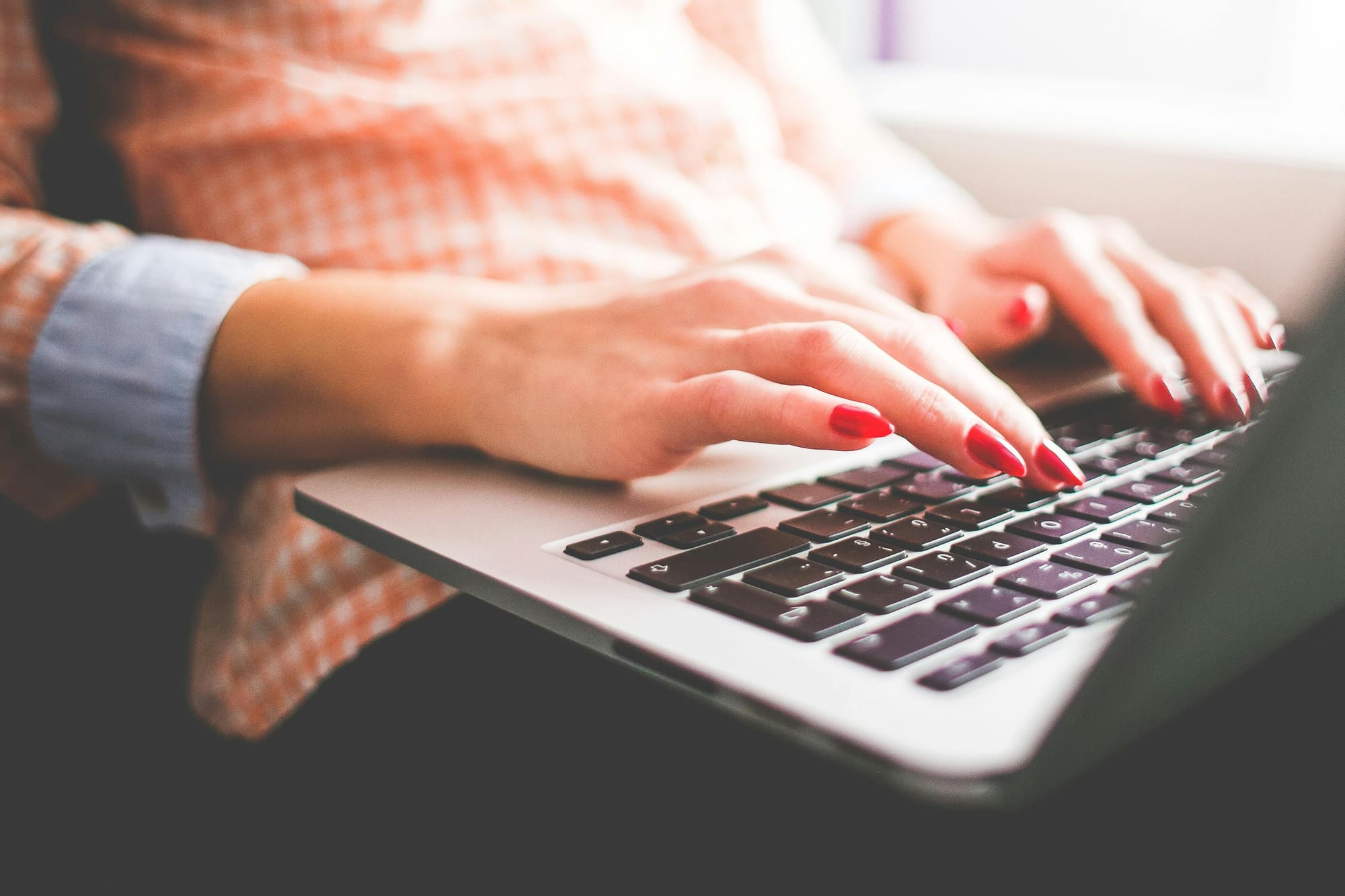 A close-up of a woman’s hands with red nail polish typing on a laptop keyboard.